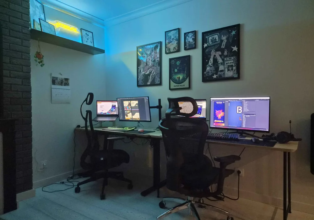 The studio's office: two workstations in bluish light, facing a wall of framed illustrations